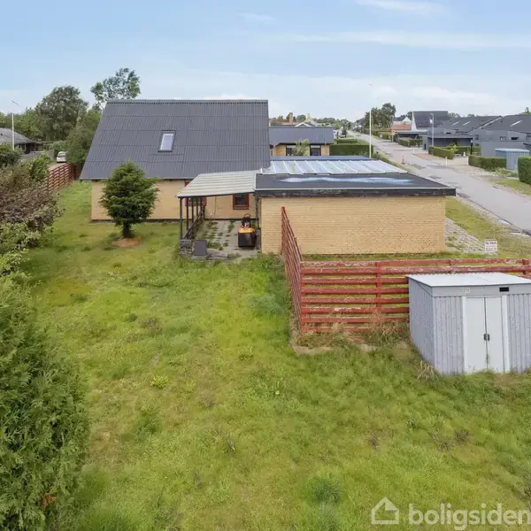 En gul murstensvilla med gråt tag og tilhørende skur står i en frodig have med et træ. Huset ligger i et villakvarter langs en vej med flere lignende ejendomme.