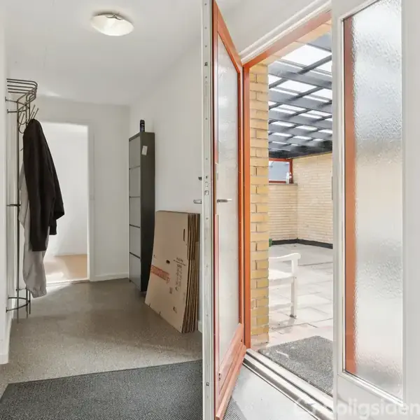 En entre med jakker hængende på en knagerække. Dør åbner til en gårdhave med fliser og murstensvægge. Kasser står op ad væggen.