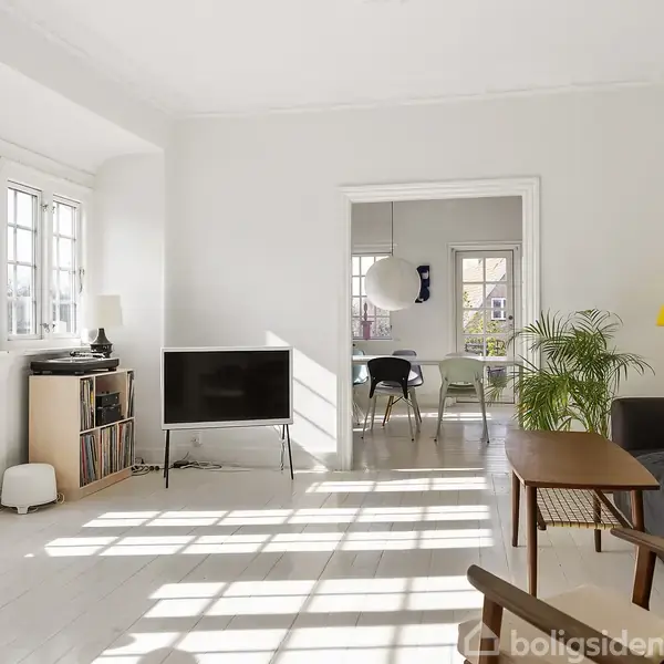 Stue med tv placeret på stativ, omgivet af lys indretning, sofa og gulvlampe. Store vinduer lader dagslys strømme ind. I baggrunden ses et spisebord med stole.