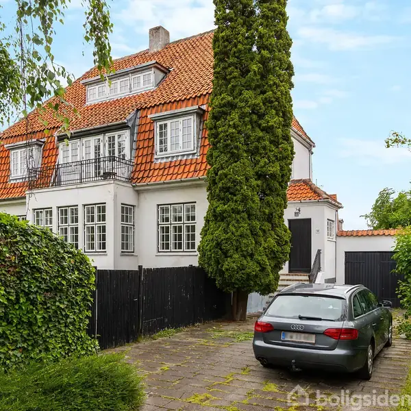 Rødt tegltag hus med flere etager, omgivet af grønne planter og træer. En grå bil holder parkeret i indkørslen ved siden af huset.