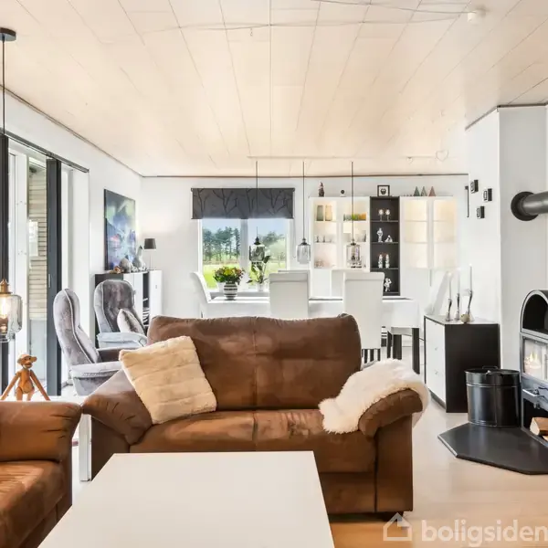 Brun lædersofa med puder står i en stue; en pejs til højre er tændt. I baggrunden ses spisebord og reoler. Store vinduer slipper lys ind i det moderne rum.