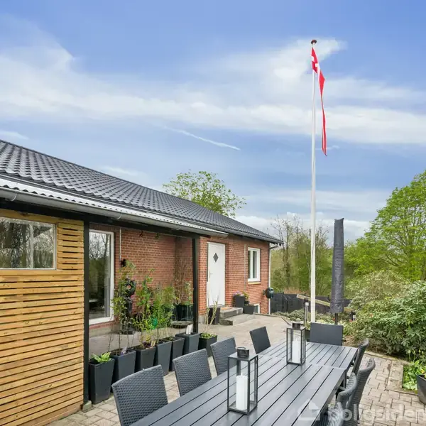 En lang sort udendørs spisebord med lanterner står på en terrasse ved siden af et murstenshus. Der er potteplanter langs væggen, og et dansk flag vajer i en flagstang.