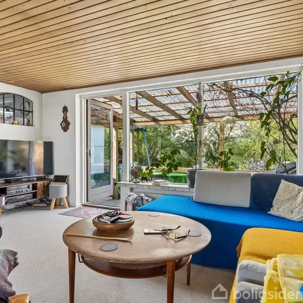 Stue med blå og gul sofa vender mod træbord med genstande. Glasdøre åbner mod overdækket terrasse med planter. Tv-stativ og dekorationer på væg. Rummeligt og lyst interiør.