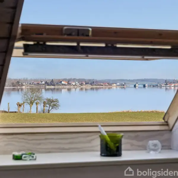 En stille sø med klart vand reflekterer den blå himmel med spredte skyer. En smal, sandet bred løber langs venstre side, mens fjerne huse ses i baggrunden.