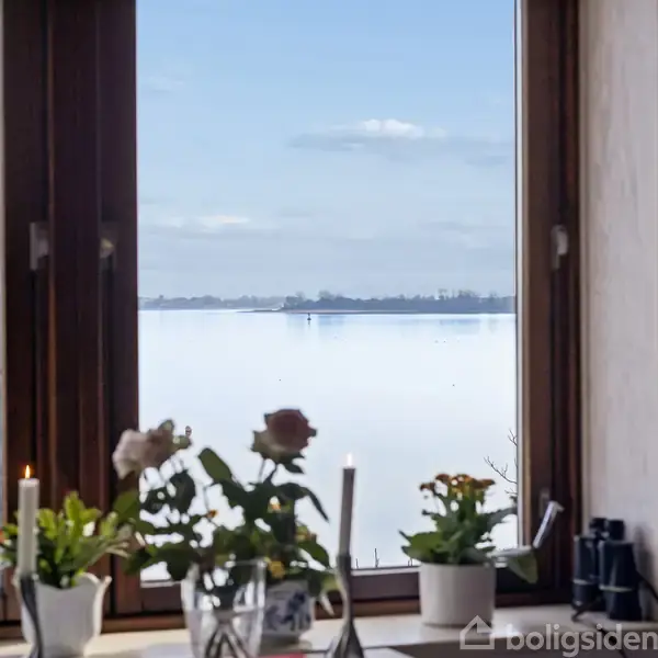 Hvid bygning med solpaneler på taget, beliggende ved kysten med en grøn have foran. Andre huse og havet ses i baggrunden under en blå himmel med skyer.