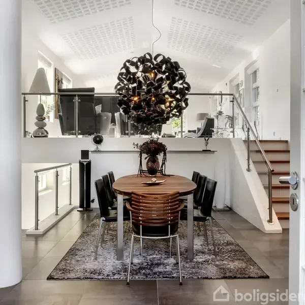 Et sort gitter indkapsler en indkørsel der fører til et moderne hus med gråt tag, omkranset af velplejede træer og buske. Et skilt viser tallet "60".