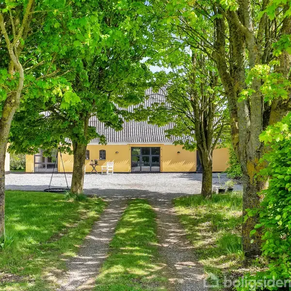En gul bygning står stille bag en træomkranset indkørsel med en lille sving og en stol foran. Grønne blade skaber en frodig ramme.