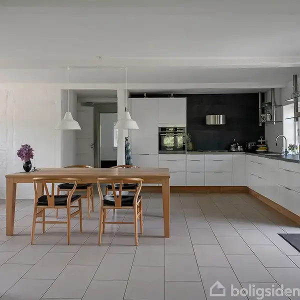 Et køkken med hvide skabe og en sort væg har et langbord med fire stole i midten. Vinduer og køkkenudstyr skaber en moderne atmosfære. Indretningen er minimalistisk og lys.
