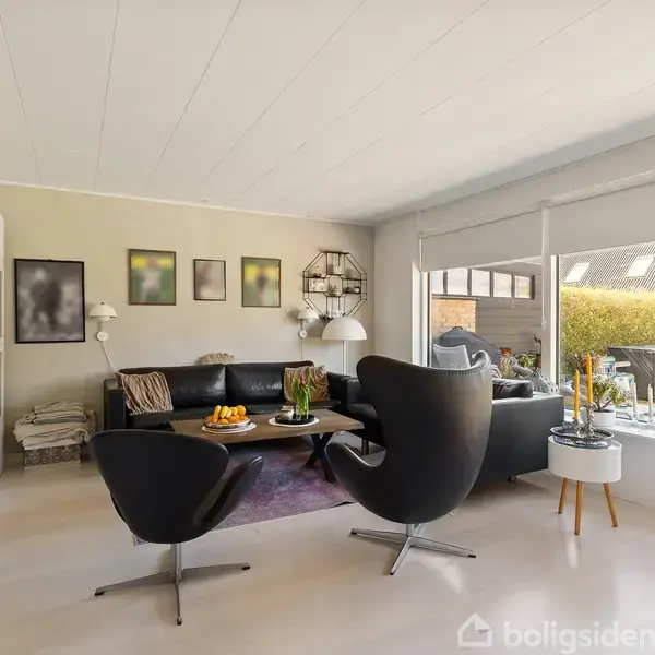 To lænestole og en sofa står i stue, hvor en sofabord med frugtskål er foran. Store vinduer giver udsigt til en terrasse. Vægge har forskellige kunstværker.