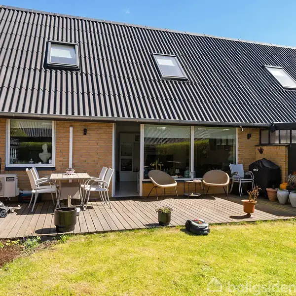 Rækkehus med terrasse møbleret med borde og stole. Foran er en veltrimmet græsplæne og krukker med planter. Terrassen grænser op til en murstensmur med store vinduer.