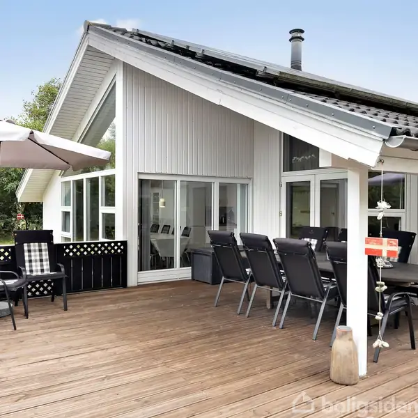 Træterrasse med hvidt sommerhus i baggrunden. Havemøbler står på terrassen, herunder et sæt med parasol og et spisebord med stole. Omgivet af grønne træer.