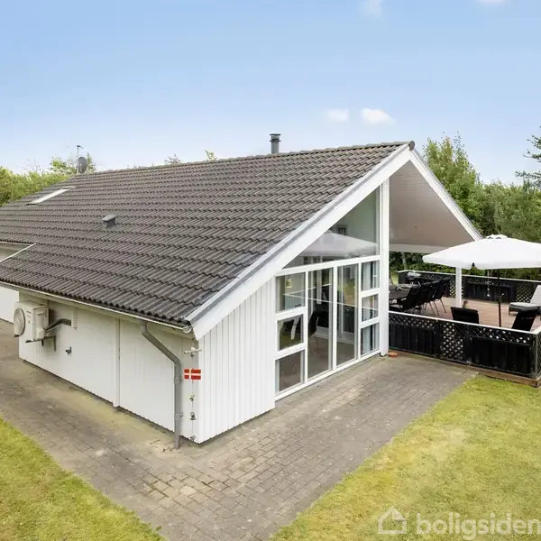 En hvid, moderne sommerhus med store vinduespartier står stille. Ved siden af er der en terrasse med udendørsmøbler og en parasol. Omgivet af en græsplæne og træer.