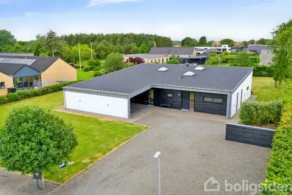 Et moderne, sort-hvidt hus står stille i en forstad, omgivet af velplejede græsplæner og hække. Flere huse og træer ses i baggrunden under en blå himmel.