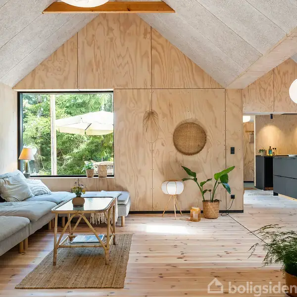 En stue med en grå sofa og sofabord, omgivet af lyse trægulve og vægge. Store vinduer viser en frodig grøn have udenfor. Roligt, minimalistisk interiør med planter.