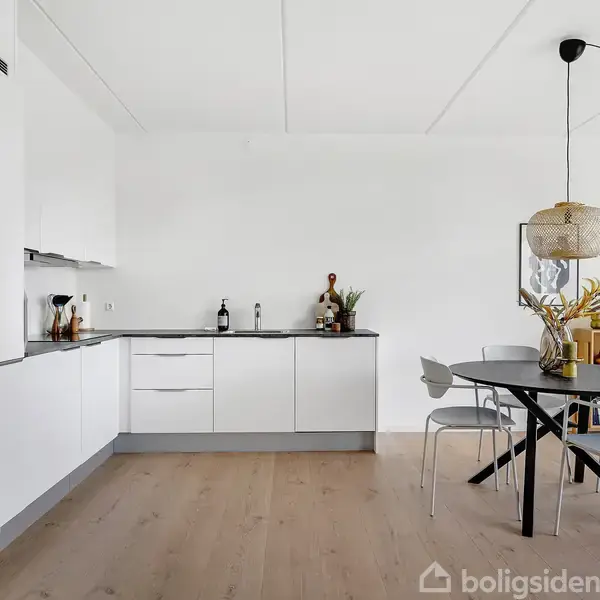 Et hvidt køkken med moderne skabe og indbygget ovn; et rundt spisebord med fire stole står i rummet, dekoreret med en lampe og planter på bordet og reolen.