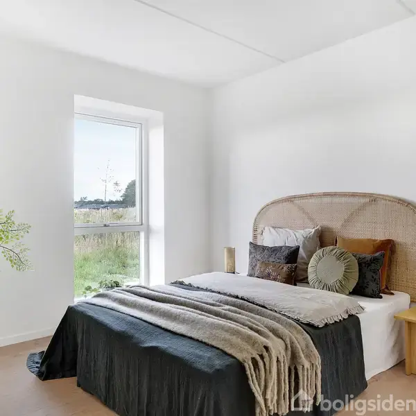 Seng i lyst soveværelse med flet-gavl har puder og tæpper, står ved vindue med udsigt til natur. En lille træhylde med lys ved siden og en grøn plante til venstre.