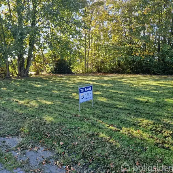 En græsplæne med et skilt placeret i midten. Skiltet viser teksten: "TIL SALG" omgivet af grønne træer i baggrunden under en solrig himmel.