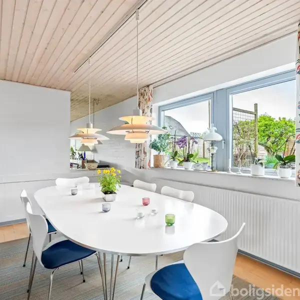 Langt, hvidt spisebord med blåstole omkring, står under moderne lamper. Lyst rum med træloft, stort vindue og udsigt til have. Grønne planter på vindueskarmen skaber en naturlig atmosfære.