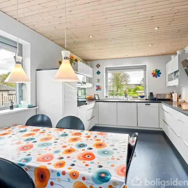 Et moderne køkken med hvide skabe og skuffer. Der er et spisebord med en farverig blomsterdug og sorte stole. Store vinduer giver udsigt til en have.