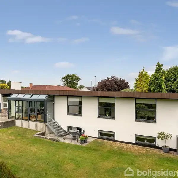 En moderne villa med store vinduer står stille i en velplejet have med grønne buske og træer, der omkranser huset. Terrasse til højre for dobbeltdøre.