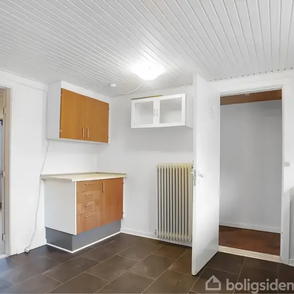 Et køkken med hvide vægge og træloft. Køkkenskabe er orangebrune, og der er en radiator under et åbent skab. En dør fører til et tilstødende rum.