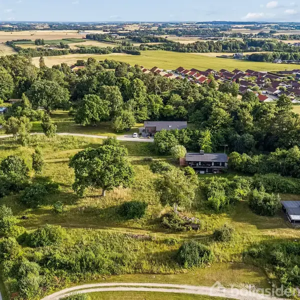 En skovklædt landskab med spredte huse og en snoet vej. Husene ligger i en grøn, landlig setting med marker og bebyggelse i baggrunden under en blå himmel.