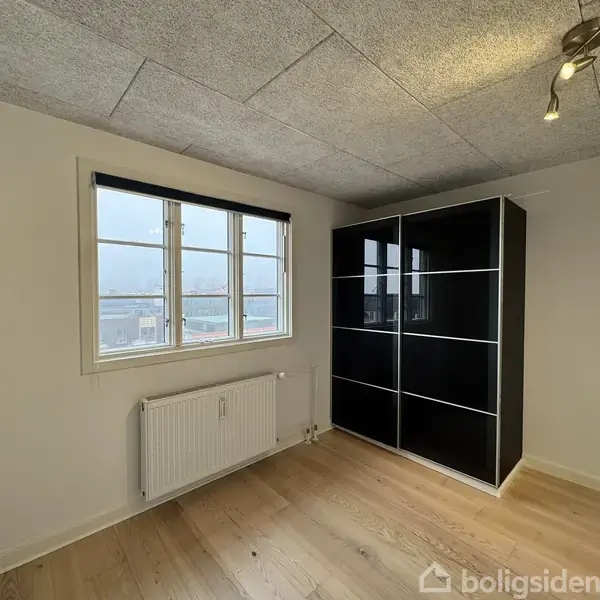 Et værelse indeholder et sort skab med hvide striber, står ved en hvid væg. En radiator under et vindue med udsigt til bygninger, og loftet har indbygget belysning.