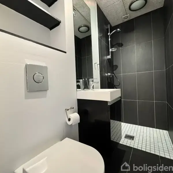 Toilet med trykknap hænger på en hvid væg, ved siden af en papirholder. Håndvask står under et spejl, og der er en brusekabine med sorte fliser i baggrunden.