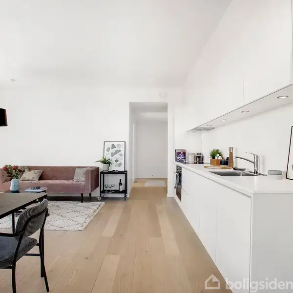 Stue med moderne indretning, hvor en hvid køkkensektion er integreret. Stue med sofa, tæppe og lamper. Et rundt spisebord står ved siden af køkkenet med stole omkring.