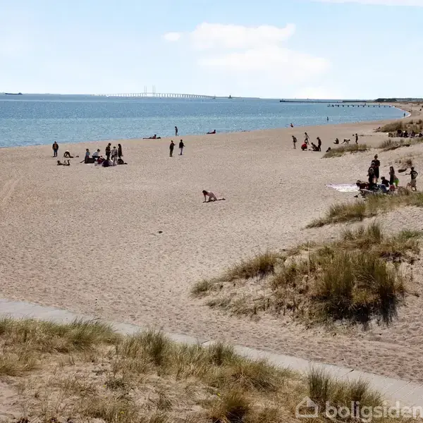 Strand med sand og græsbevoksning, hvor flere grupper af mennesker slapper af og nyder solen ved vandet. En bro ses i baggrunden hen over horisonten.