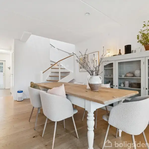 Et lyst spisebord med seks stole står i et moderne køkkenalrum. Bordet er dekoreret med en vase med grene. I baggrunden ses en hvid trappe og en skænk med planter.