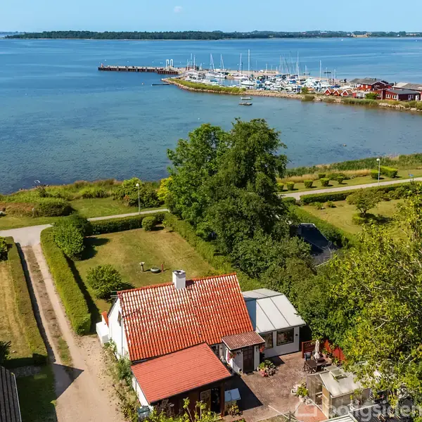 Et hus med rødt tegltag ligger ved vandet i en naturskøn kystlandsby. Foran huset er en grusvej, mens en marina med både ses i baggrunden.