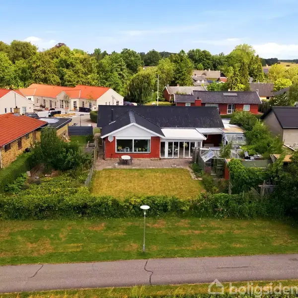 Hus i forgrunden med rød murstensfacade og stor have, omgivet af grønne buske. Flere huse i baggrunden og skov i horisonten under en blå himmel.