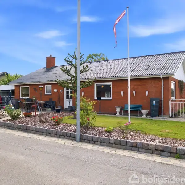 Et murstenshus står stille i en villavej. Huset har et gråt tegltag, flagstang med flag, og er omgivet af en velholdt have. Et skilt i haven lyder "home til salg".