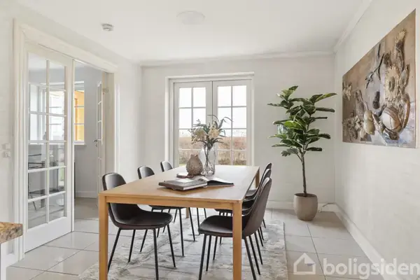 En gul murstensvilla med rødt tegltag står bag en grusplads omgivet af grønne træer, nær en strand. En hvid port åbner ud til en vej.