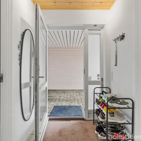 Gangen har en skohylde fyldt med sko ved siden af en vaskemaskine og tørretumbler. Der er et stort spejl på væggen og en åben dør fører ud til en overdækket veranda.
