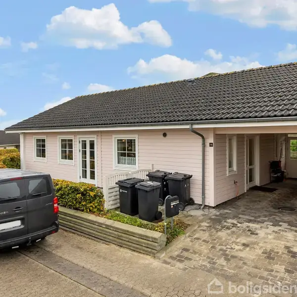 En grå bil står parkeret foran et lyserødt hus med sort tag. Huset har en carport til højre og flere vinduer. En hæk omkranser ejendommen.