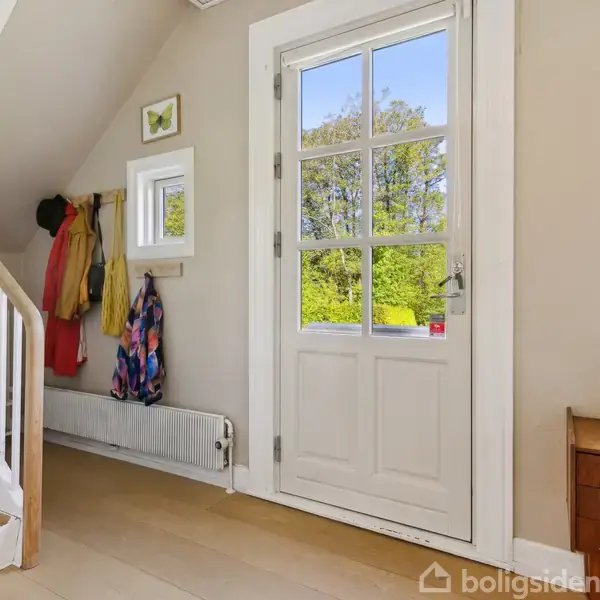 En entre med hvid dør med glas, ved siden af en stige op ad væggen. Jakker hænger på knager over en radiator. Sollys trænger ind gennem vinduet.