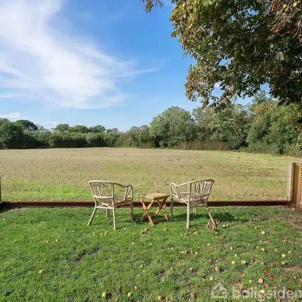 To stole står ved et lille havebord på en græsplæne, omgivet af hegn; i baggrunden ses en åben mark og træer.