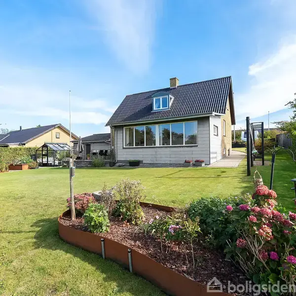 Et hvidt hus med sort tag står i en velplejet have med blomsterbed i forgrunden. Der er blå himmel og græsplæne omkranset af hække.