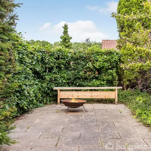 Bålsted med flammer står på en flisebelagt terrasse foran en træbænk. Omgivet af tætte grønne buske og træer i en have.