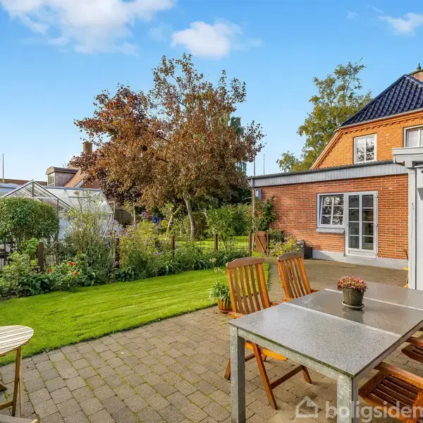 Terrassebord med stole står på en flisebelagt patio. I baggrunden ses en have med græsplæne, planter, et drivhus og et rødt hus med tag af teglsten.