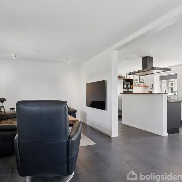 Sort læderstol står i stue, vendt mod mørk sofa. Stuen åbner op til et moderne køkken med hvide skabe og fritstående emhætte. Væggene er hvide, og gulvet er mørkt.