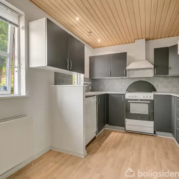Køkken med mørke skabe og en hvidevareovn er ubenyttet. Det har trægulv og loft samt et vindue med udsigt til træer udenfor.