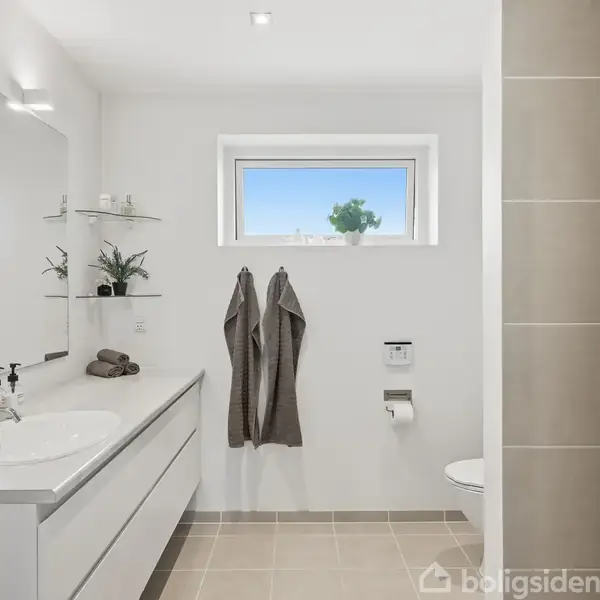 Et badeværelse med en moderne stil. En hvid vask med skuffer er placeret til venstre, to håndklæder hænger på væggen, og et lille vindue giver udsigt til himlen.
