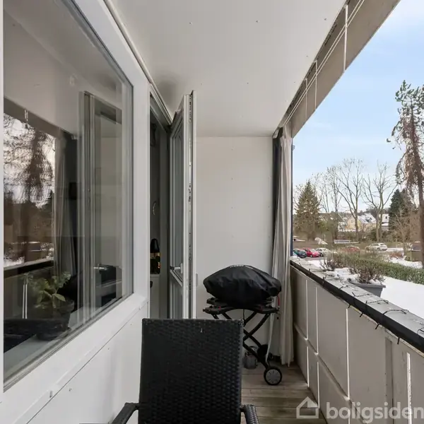 En altan med en grill under et halvtag ved siden af en stol. I baggrunden ses træer og parkerede biler i et beboelsesområde.