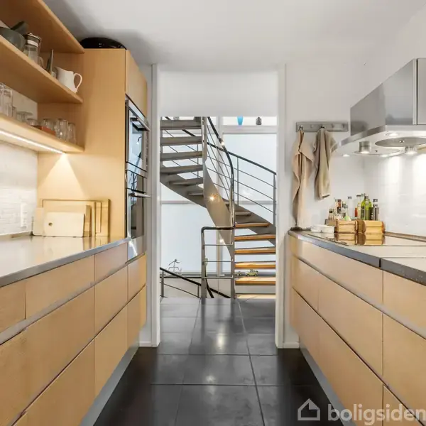 Et moderne køkken med træskabe og sorte bordplader står på begge sider af et smalt rum. I baggrunden ses en vindeltrappe, mens der er glas og tallerkener på hylderne.