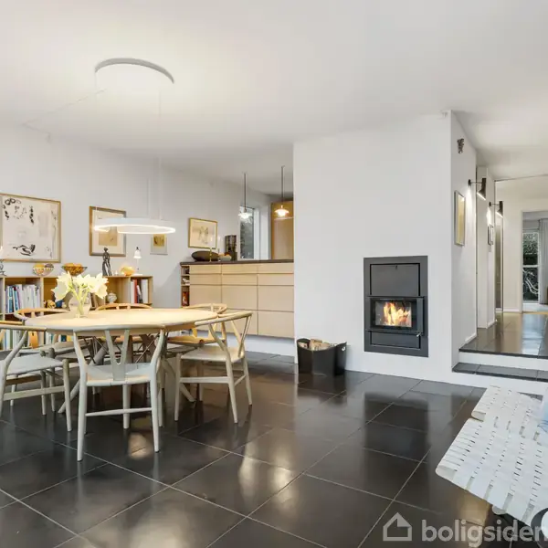 Spisebord omgivet af stole står midt i et rum med sort flisegulv. Ved siden er en bogreol, billeder hænger på væggen, og en brændeovn er indbygget i væggen.
