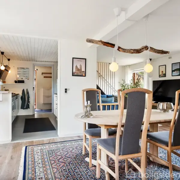 Spisebord med seks stole står på et mønstret tæppe i en stue. I baggrunden ses et åbent køkken med barstol, og et stort vindue slipper lys ind.