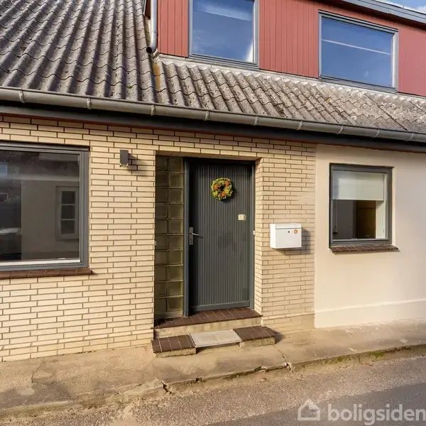 Et rækkehus med en grå hoveddør dekoreret med en krans, placeret mellem to vinduer. Konteksten viser en rolig gade med en murstensfacade.